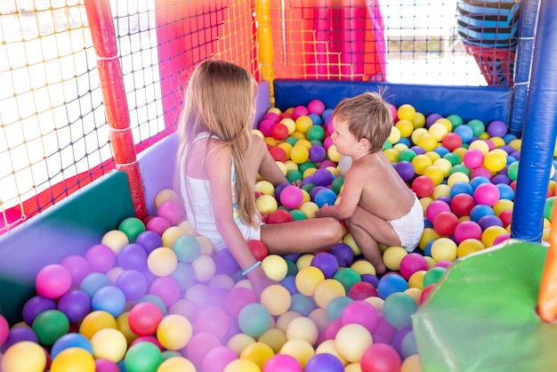 Cheerful beautiful bright older sister is playing with her cute little cute little brother and throws colorful little balls at him while sitting in a small playroom