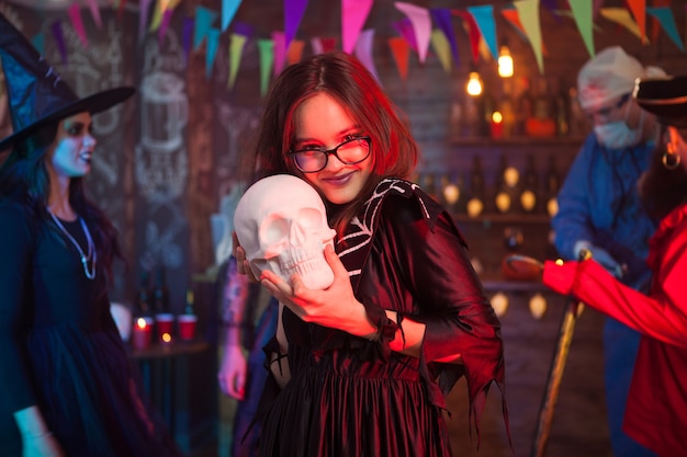 Photo cheerful and beautiful girl holding a human skull for halloween celebration. girl in witch costume.