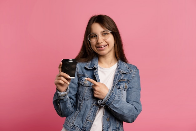 Cheerful beautiful woman blogger in glasses, holds take way cup and points to the cup a finger