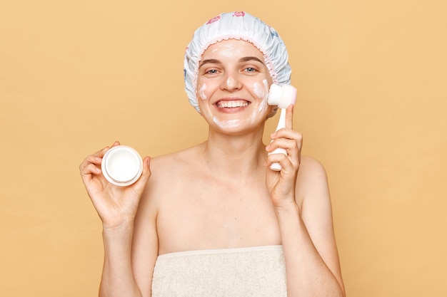 Cheerful beautiful woman doing skin care procedures cleaning her face with brush holding cream in hands looking at camera with satisfied expression