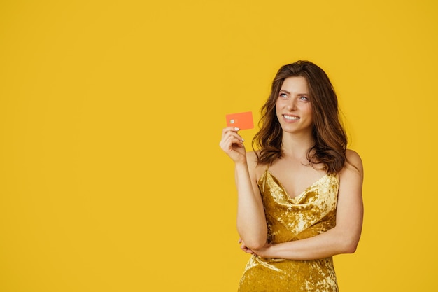 Cheerful beautiful woman in elegant dress holding credit card and dreaming about buying yellow