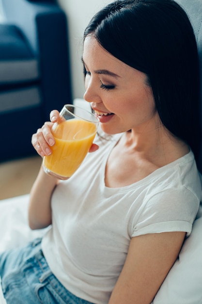 Cheerful beautiful young lady sitting at home and looking happy while drinking orange juice from the glass