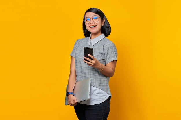 Cheerful beautiful young woman looking at mobile phone screen and holding laptop on yellow background