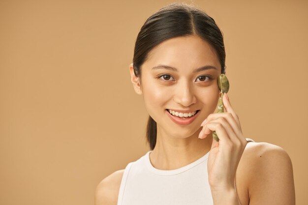 Cheerful beautiful young woman smiling at camera while using jade roller for massaging her face