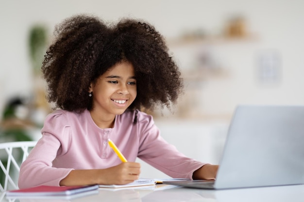 Cheerful black girl schooler doing homework using laptop