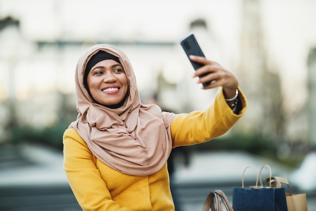 Cheerful black Muslim woman wearing a hijab and making a selfie by her smartphone in urban environment.