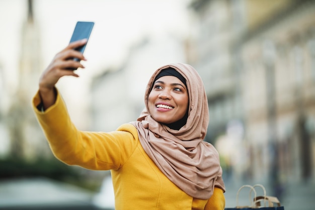 Cheerful black Muslim woman wearing a hijab and making a selfie by her smartphone in urban environment.