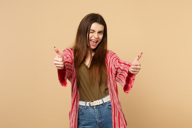 Cheerful blinking young woman in casual clothes looking camera, showing thumbs up isolated on pastel beige wall background in studio. People sincere emotions, lifestyle concept. Mock up copy space.