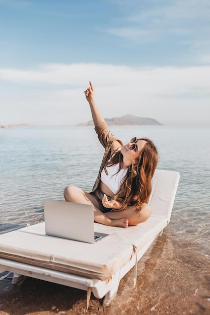 A cheerful blogger girl remotely works with a laptop on the beach with sea views A woman makes a video from a trip Travels the world and works with social networks online Remote modern profession