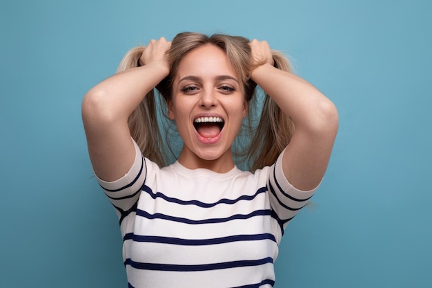 Cheerful blond young woman holds her hair in her hands and laughs cutely on a blue background