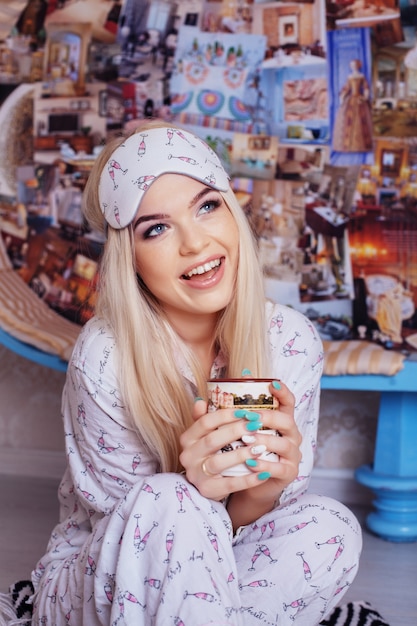 Cheerful blonde girl drinks coffee in pajamas. Sleep mask. Concept lifestyle, rest, breakfast, sleep.