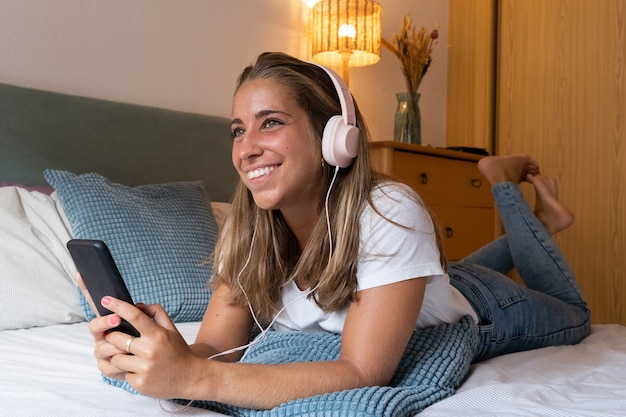 Cheerful blonde woman using smartphone indoors. Horizontal side view of caucasian woman listening to music with headphones lying in bed at home. Technology and women lifestyle indoors