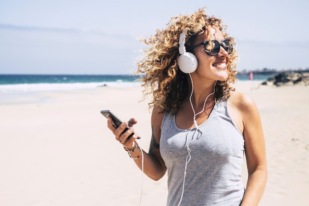 Cheerful blonde woman with curly hair enjoying the summer on the beach listening to music