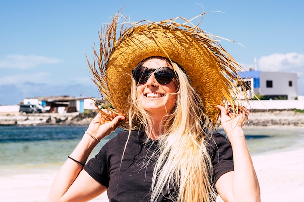 Cheerful blonde young girl with tourist style hat under the sun in a great day of outdoor vacation on the beach