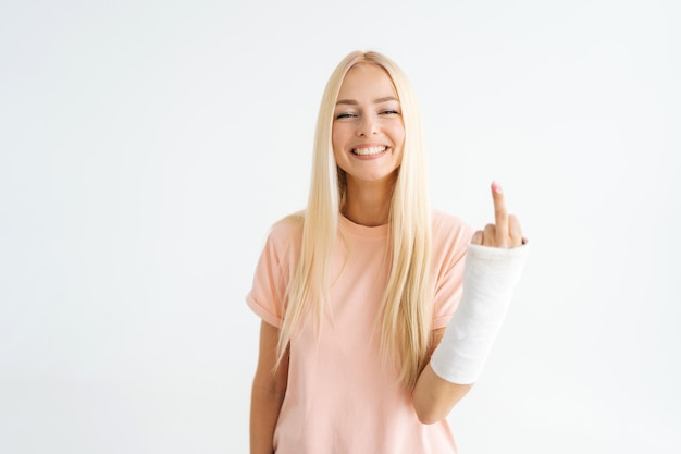 Cheerful blonde young woman showing middle finger rude gesture with broken arm wrapped in plaster bandage standing on white isolated background