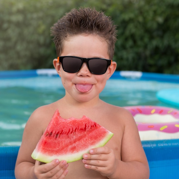 Cheerful boy by the blue pool holding a slice of ripe bitten watermelon sticking out his tongue concept