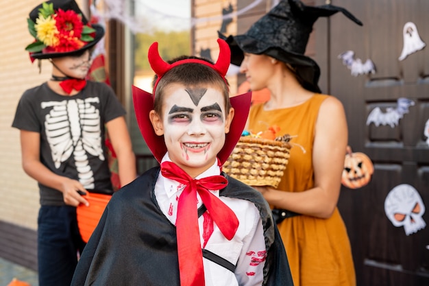 Cheerful boy in halloween costume looking at you against young woman in yellow dress and hat holding basket with treats and his friend