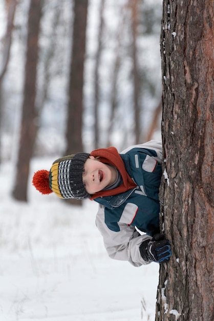 Cheerful boy peeks out from behind tree Child in knitted hat walks through winter snowkept park