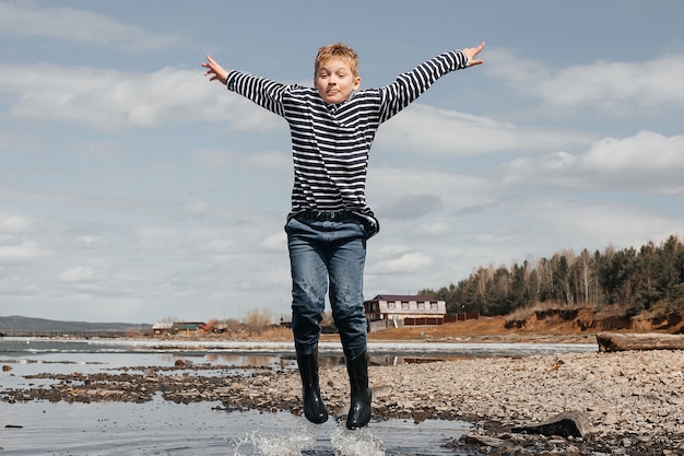 A cheerful boy in a striped vest and rubber boots jumps in the water on the river bank.