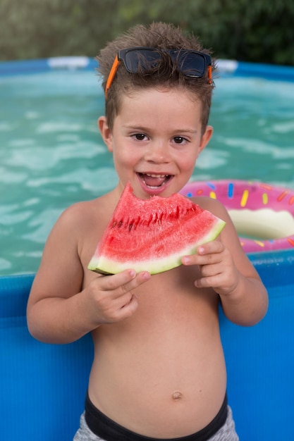 Cheerful boy with glasses on his forehead eating ripe watermelon against the background of a street pool concept