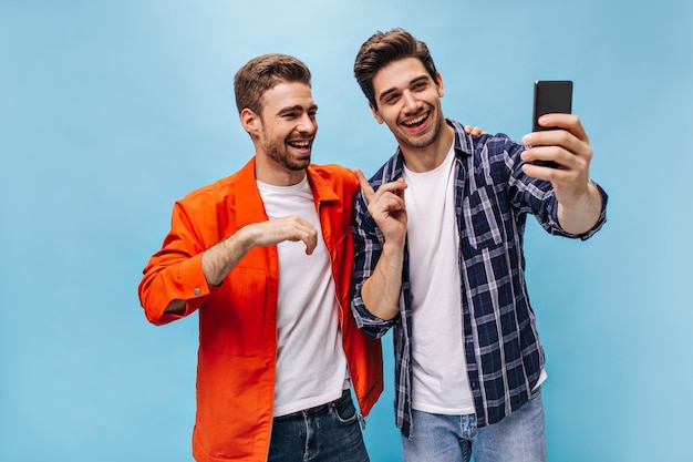 Cheerful brunet men in jeans smile and take selfie in great mood on blue background Guy in checkered shirt holds phone