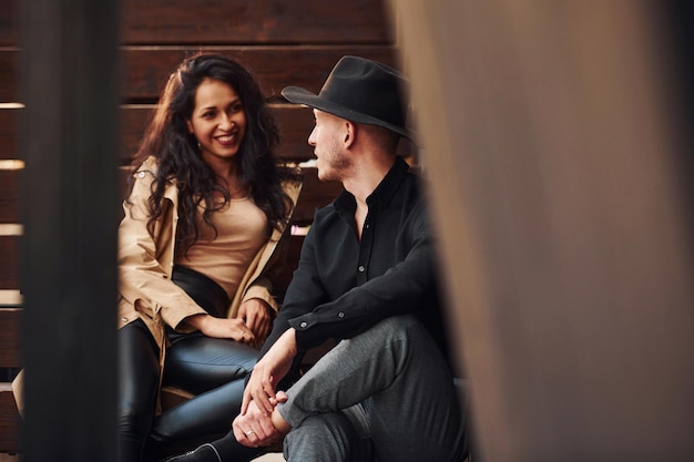 Cheerful brunette in black leggings sitting on wooden stairs with her man in hat