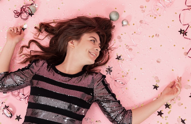 Cheerful brunette girl lies on a pink background among confetti and streamer, the concept of celebrating christmas and new year, festive party.