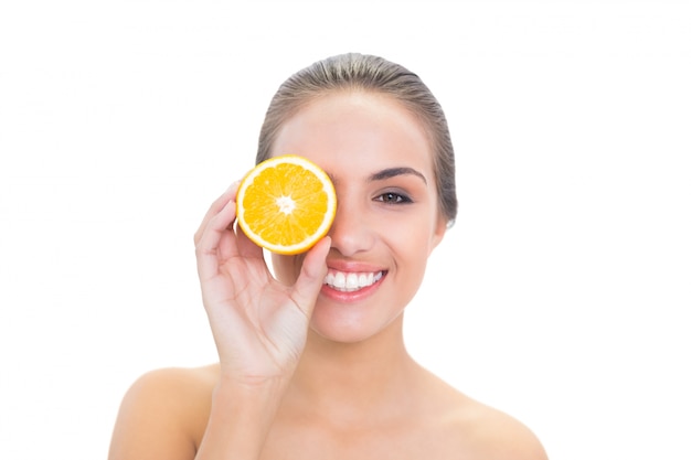 Cheerful brunette woman holding an orange half in front of her eye