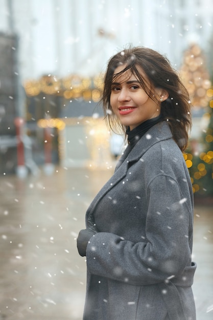 Cheerful brunette woman wears trendy coat walking at the city decorated to Christmas during the snowfall