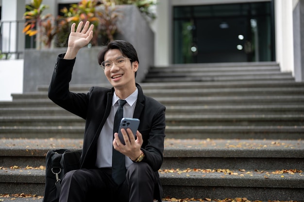 A cheerful businessman is waving his hand to say hi to his colleagues while sitting on the stairs