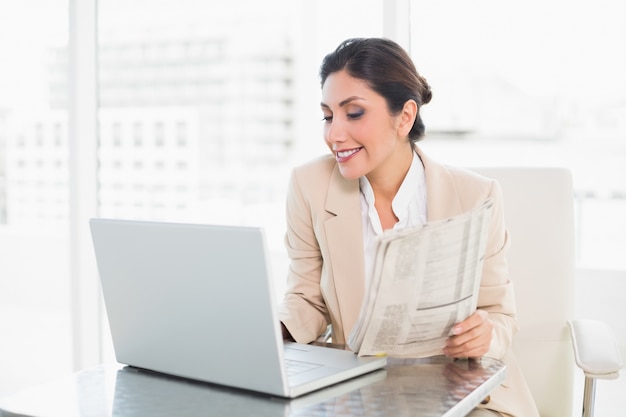 Cheerful businesswoman holding newspaper while working on laptop