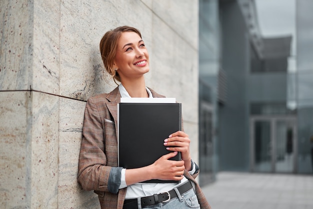 Cheerful businesswoman leaning at building office, holding documents, enjoying break outdoors