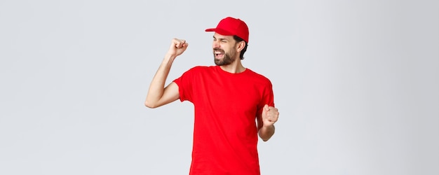 Cheerful and carefree young bearded delivery guy in red uniform cap and tshirt singing yelling yes and fist pump in celebration triumphing and rejoicing over great news grey background