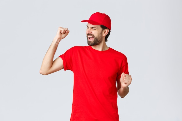 Cheerful and carefree young bearded delivery guy in red uniform cap and tshirt singing yelling yes a...