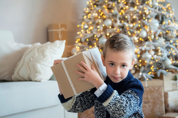 Cheerful Caucasian boy raising wrapped gift, trying guess what is inside, feeling excited opening New Year presents. Little child checking Christmas presents.