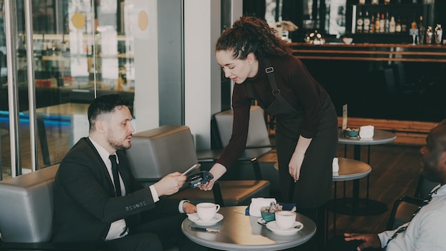 Cheerful Caucasian businessman sitting with his African American partner and paying online bill using his smartphone in modern cafe Young waitress using cash terminal