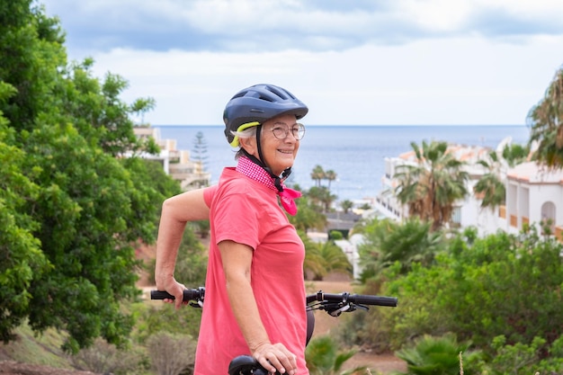 Cheerful caucasian cyclist senior woman in pink jersey and helmet standing on the hill in front to the sea in summer holidays