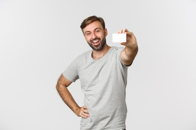 Cheerful caucasian man in gray t-shirt, showing credit card and smiling, standing over white