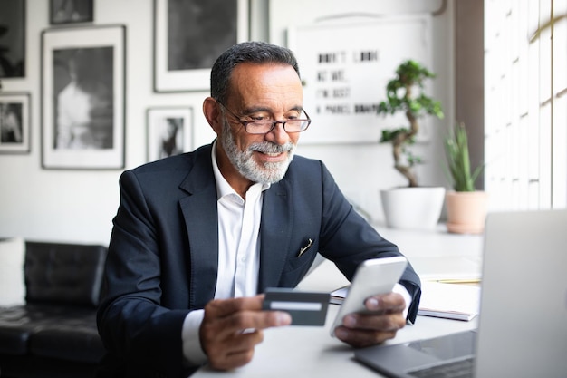 Cheerful caucasian senior businessman in suit glasses pay at phone at table with computer uses