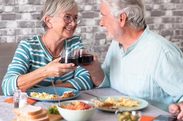 Cheerful caucasian senior couple toasting with red wineglass while sitting face to face at table having meal or brunch together at home