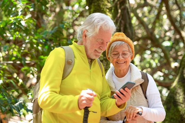 Cheerful caucasian senior couple with backpack and walking sticks hike in the forest while using mobile phone elderly couple enjoying healthy lifestyle and retirement