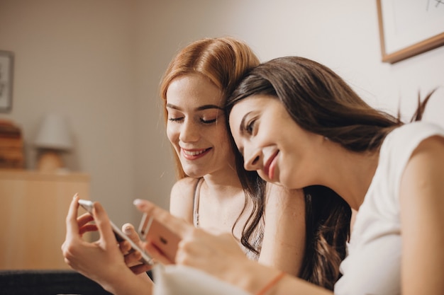 Cheerful caucasian woman with ginger hair is showing something to her brunette friend on phone while sitting on the couch