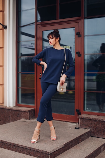 Cheerful caucasian woman with long dark hair stands near the office building and shows different poses for the camera