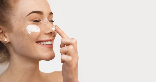 Cheerful caucasian woman with red hair and freckles is using a cream on her nose and cheeks after spa procedures