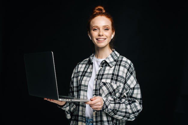 Cheerful charming young business woman or student holding laptop computer and looking at camera on isolated black background Pretty redhead lady model emotionally showing facial expressions