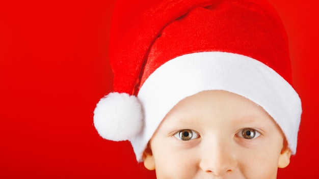 Cheerful child in a Santa hat on a red background looks out from the bottom of the frame