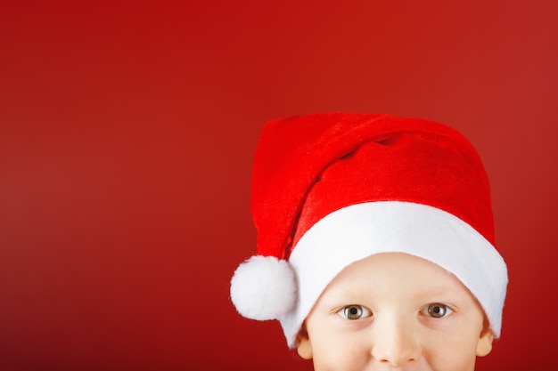 Cheerful child in a Santa hat on a red background looks out from the bottom of the frame