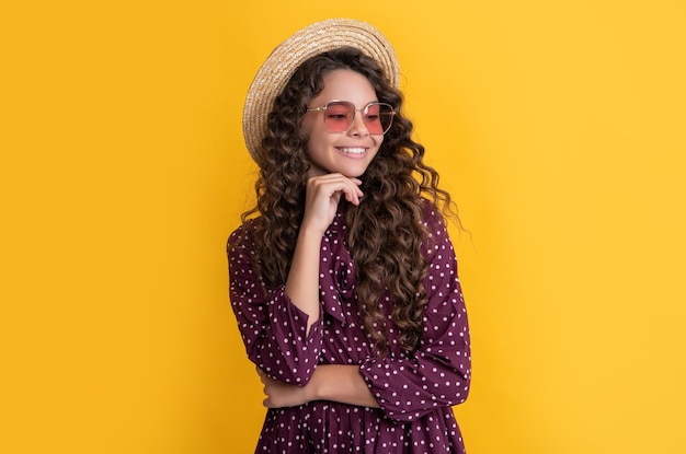 Cheerful child in straw hat and sunglasses with long brunette curly hair on yellow background