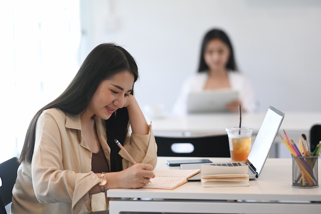 Cheerful chubby young woman sitting at desk in front of open laptop computer and making notes in diary at office.