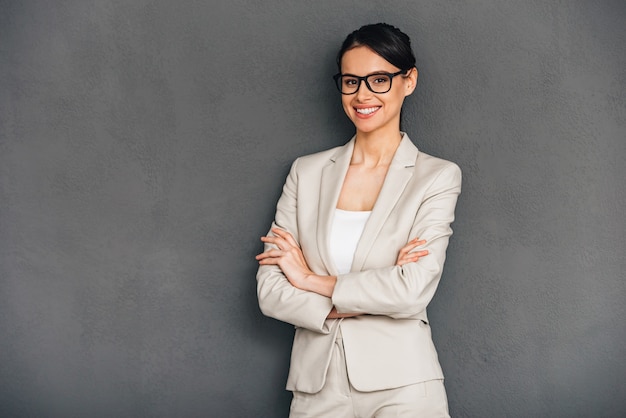 Cheerful and confidant. Cheerful young businesswoman in glasses keeping arms crossed and looking at camera with smile while standing against grey background